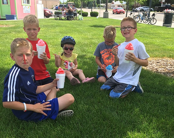 Five little kids sitting on a lawn eating shaved ice. 