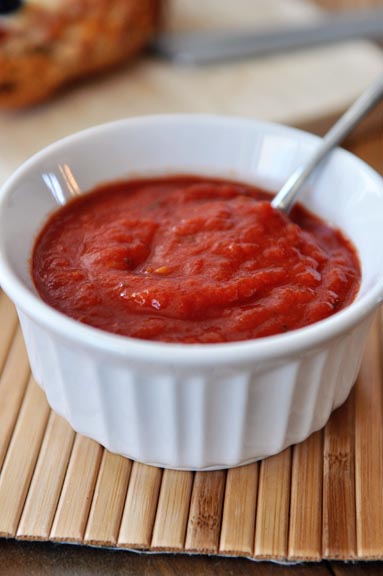 A white ramekin of red pizza sauce with a spoon in it.