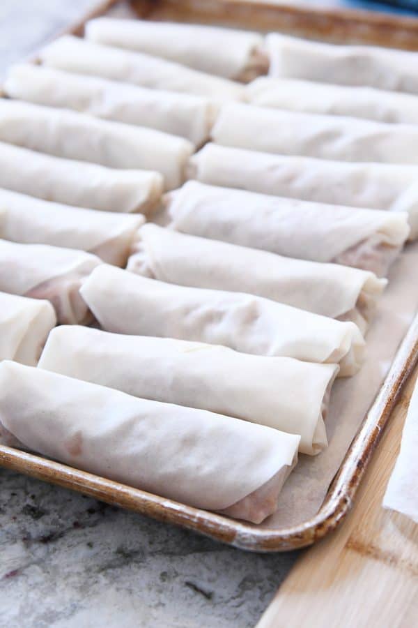 A cookie sheet full of egg rolls lined up ready to be fried.