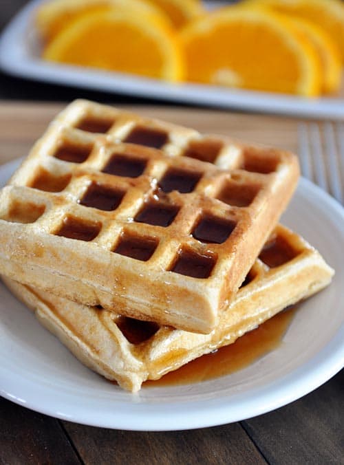Two waffles stacked on a white plate and covered with syrup with a plate of orange slices in the background.