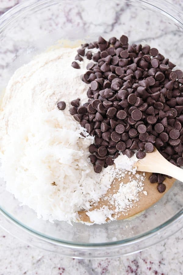 Flour, coconut and chocolate chips in glass bowl with batter ingredients.