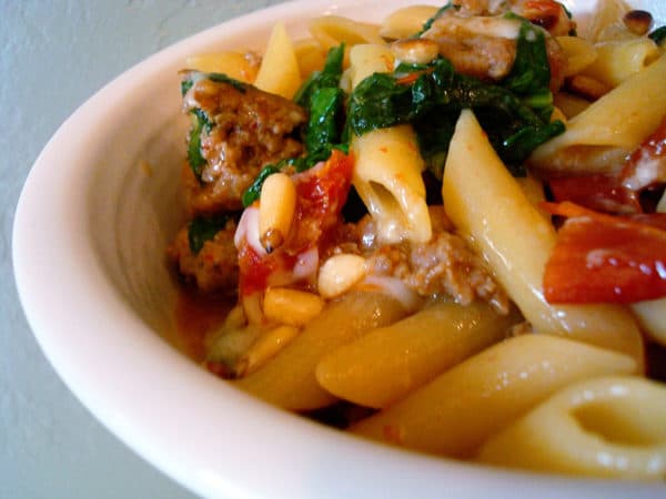 White bowl of pasta, sausage, spinach, and tomatoes.