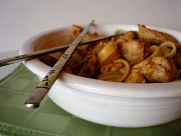 White bowl of moo shu noodles and chicken with metal chopsticks on top of the bowl.