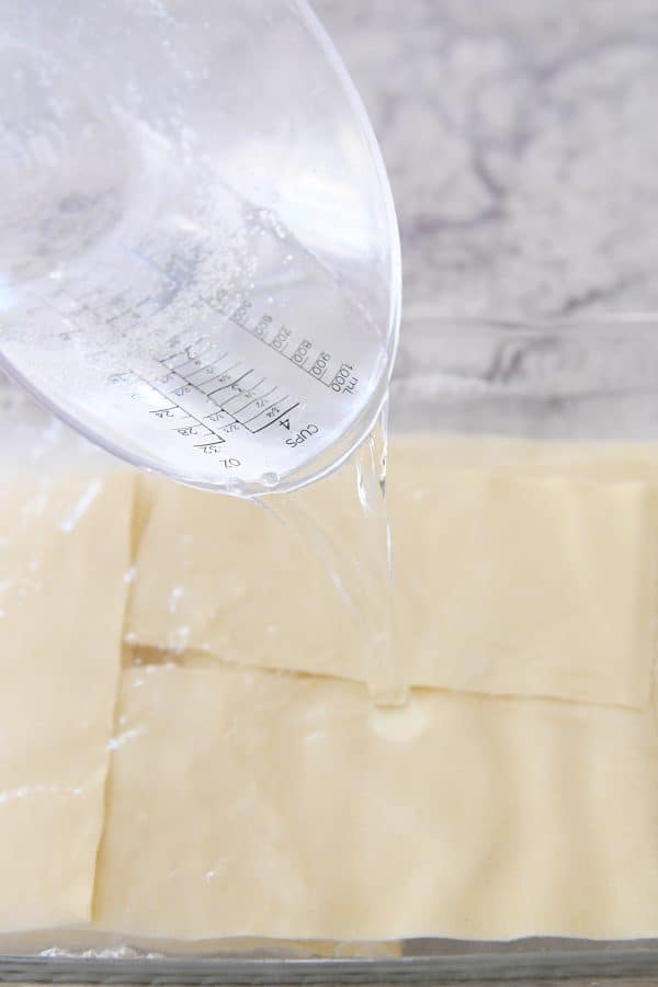 Pouring boiling water over lasagna noodles in glass pan.