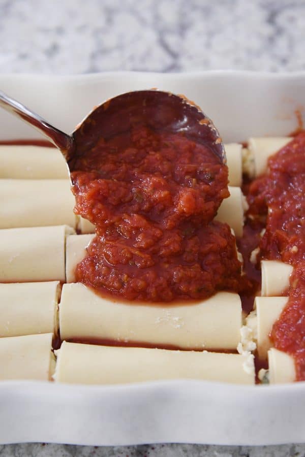 pouring red marinara sauce over rolled manicotti