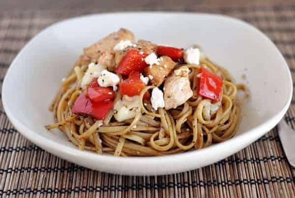 White bowl with cooked spaghetti noodles, chicken pieces, and chopped red pepper.