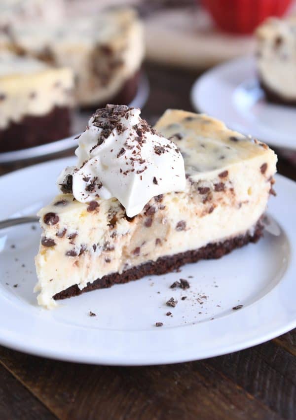 Slice of chocolate chip cookie dough cheesecake on white plate with whipped cream and chocolate shavings.