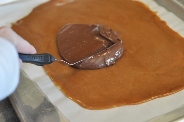 Melted chocolate being spread on top of cooled toffee on a cookie sheet.