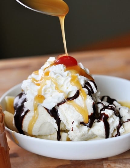 White bowl of vanilla ice cream drizzled with caramel sauce and hot fudge sauce and topped a cherry.