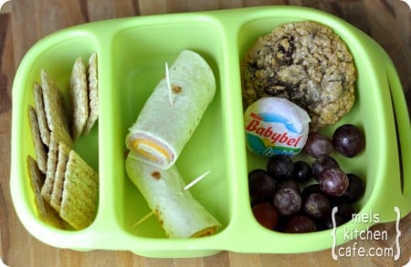 top view of a green divided lunch tray with crackers, ham cheese roll, grapes, Babybel, and cookie