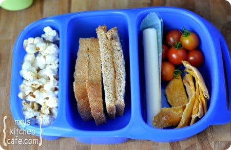 top view of a blue divided lunch box with popcorn, sandwich, cheese stick, tomatoes, and chips