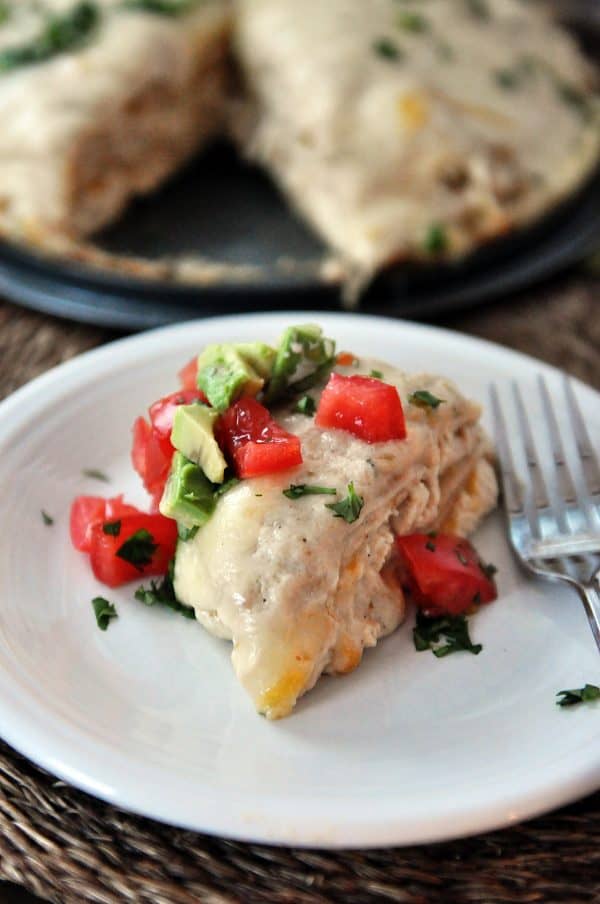 Slice of green chile chicken enchilada stack on white plate with tomatoes and cilantro and fork.