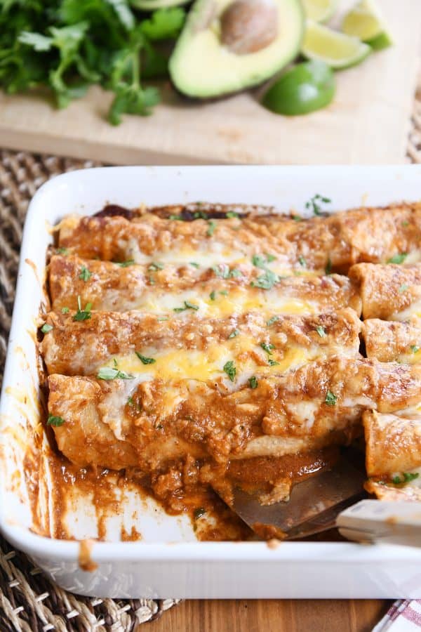 Spatula scooping out a red sauce chicken enchilada in white pan.