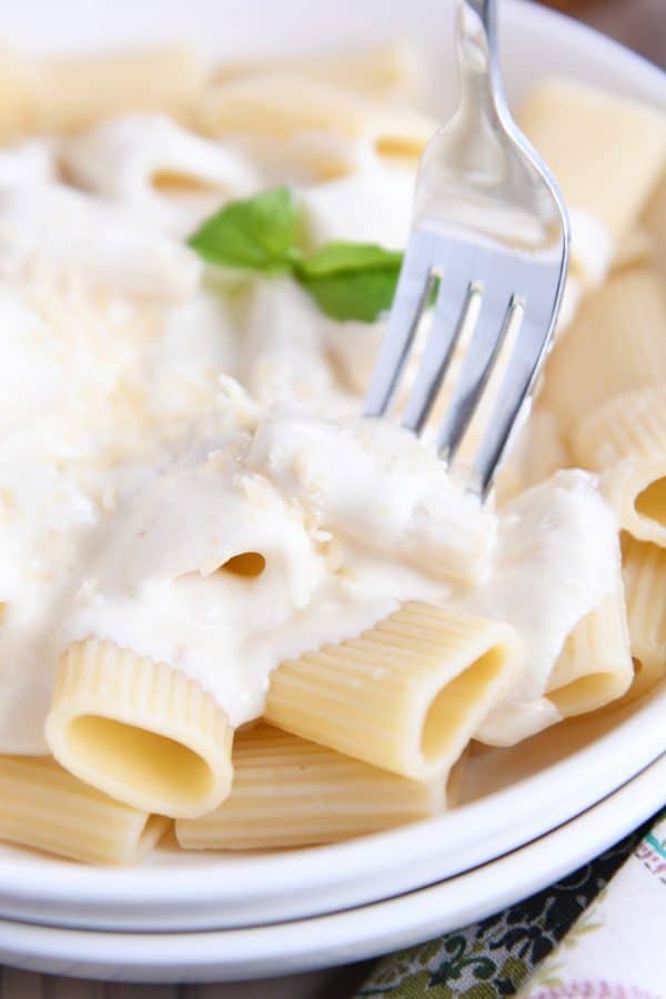 Fork taking bite out of alfredo sauce over rigatoni noodles.