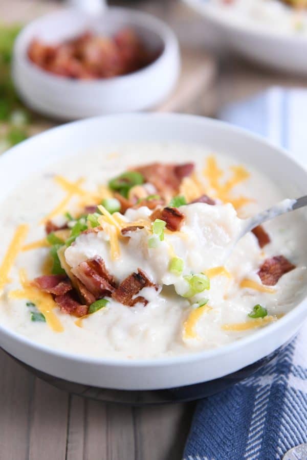Spoon taking scoop out of loaded baked potato soup in white bowl.