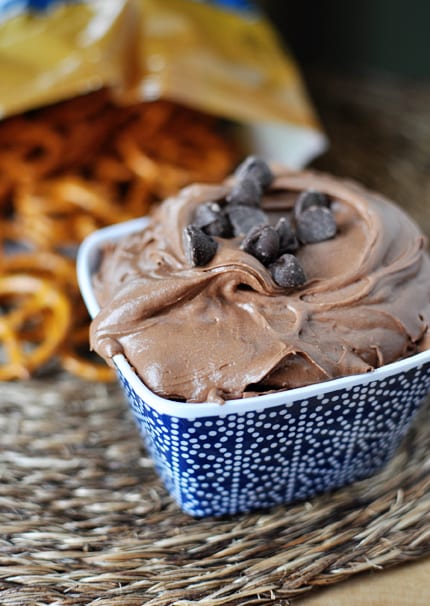 A blue ceramic bowl with chocolate brownie dip sprinkled with chocolate chips and a bag of pretzels behind it.