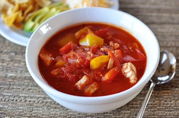 White bowl full of red chicken chili with a white plate of toppings in the background.