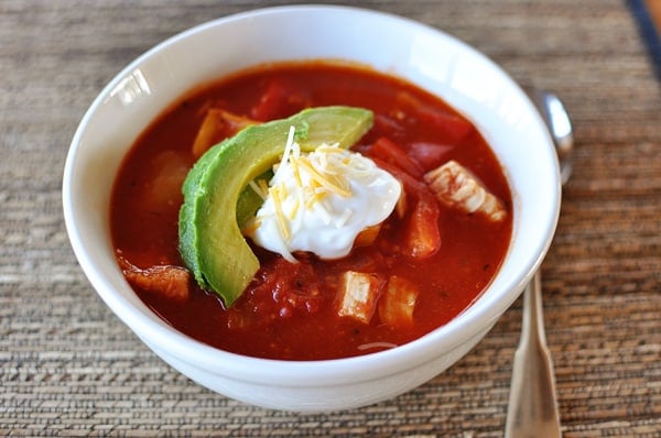 White bowl with red chicken chili topped with a slice of avocado and a dollop of sour cream.