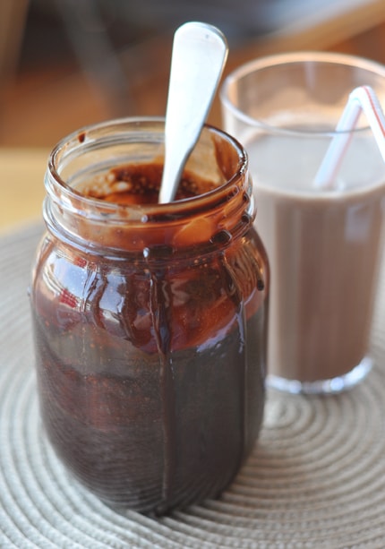 mason jar of chocolate syrup with drips coming down the side and a spoon inside in front of a glass of chocolate milk