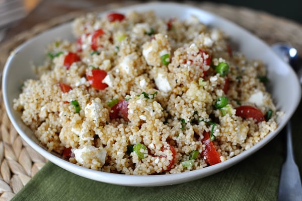 A white bowl with cooked fluffy couscous, feta, tomatoes, and peas.