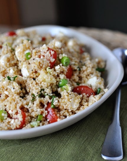 A white bowl full of cooked couscous, peas, and tomatoes.