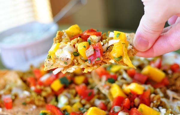 A hand holding an mango and tomato topped nacho over a full plate of nachos.