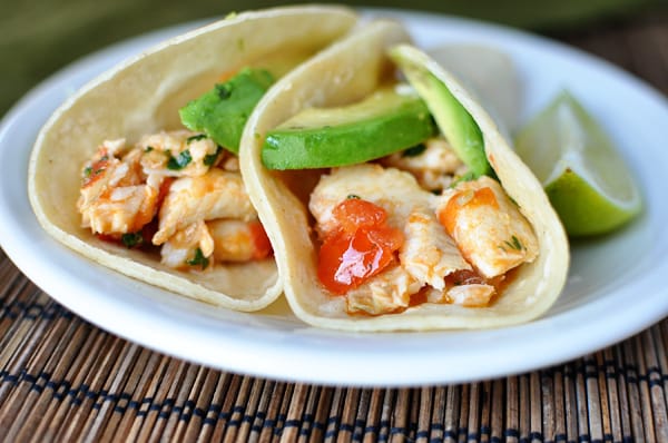 Two fish tacos in corn tortillas with avocado slices and tomatoes on a white plate.