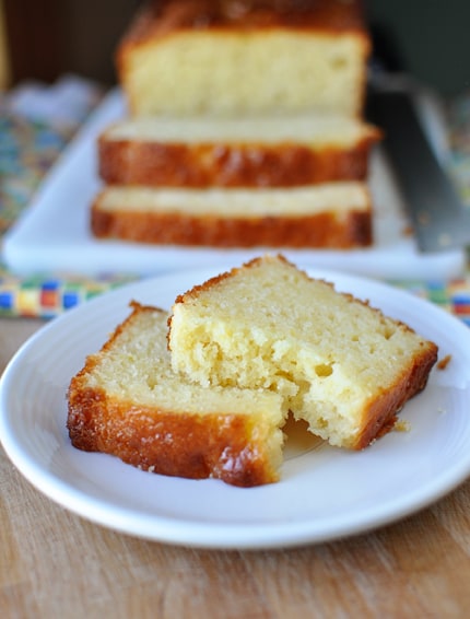A slice of lemon bread on a white plate in front of a loaf of lemon bread.