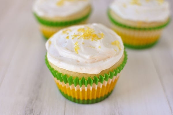 Three lemon cupcakes with white frosting and lemon zest on the top in green and yellow muffin liners.