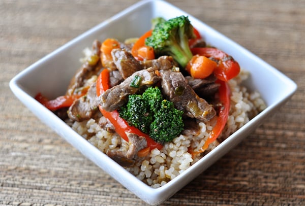 Square white bowl with cooked rice topped with cooked broccoli and peppers and beef strips.