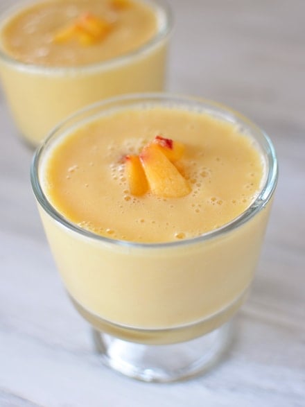Glass goblets full of peach-orange smoothie with a few diced peaches on top.