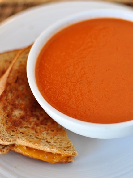 A white bowl of tomato soup with grilled cheese on the side. 