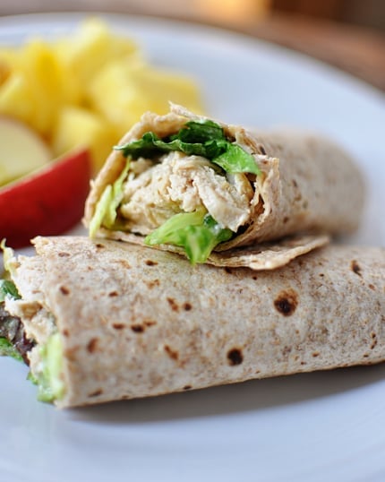 A chicken caesar salad wrap cut in half on a white plate with fruit on the background.