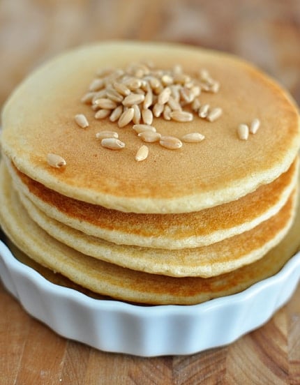 A stack of wheat pancakes in a white ramekin, with wheat berries scattered on the top pancake.