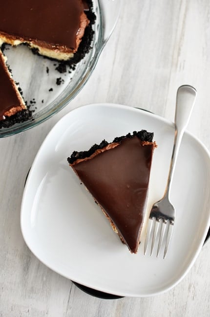 A slice of cheesecake pie with an cookie crust and chocolate ganache topping on a white plate.