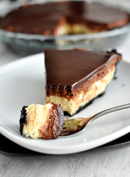 A slice of cheesecake pie covered with chocolate ganache getting a bite taken out on a white plate.
