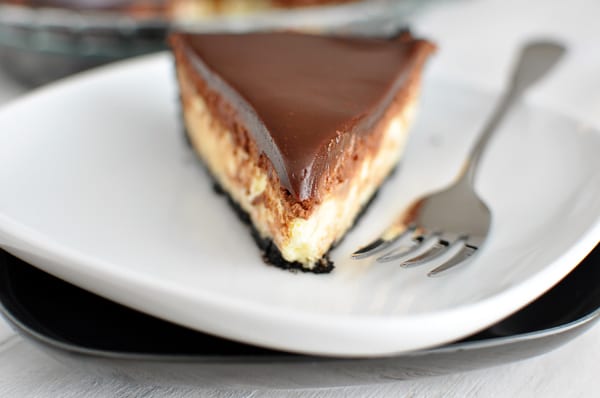 A slice of chocolate crust cheesecake pie topped with chocolate ganache on a white plate.