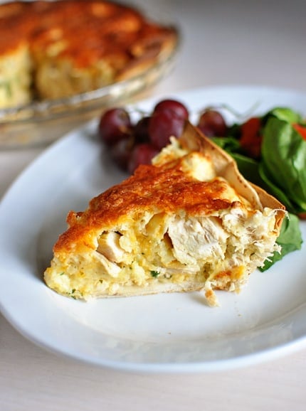 A side view of a slice of cheesy chicken quesadilla pie on a white plate next to a cluster of red grapes and green salad.