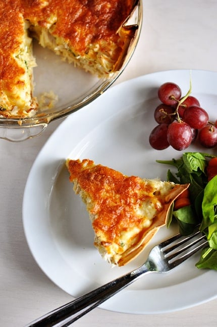 A slice of cheesy quesadilla pie on a white plate next to a cluster of grapes and green salad.