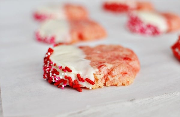 Cherry shortbread cookies half dipped in white chocolate and sprinkles laying on a sheet of parchment.