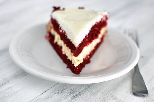 A piece of layered red velvet cheesecake cake on a white plate.