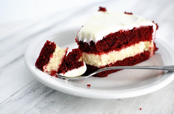 A white plate with a red velvet and cheesecake layered piece of cake with white frosting.