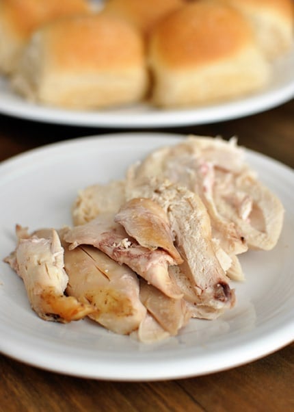 A white plate with roasted cooked chicken in front of a plate of cooked rolls.