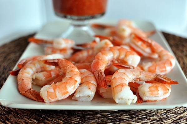 A white platter with cooked shrimp with a goblet of cocktail sauce behind them.