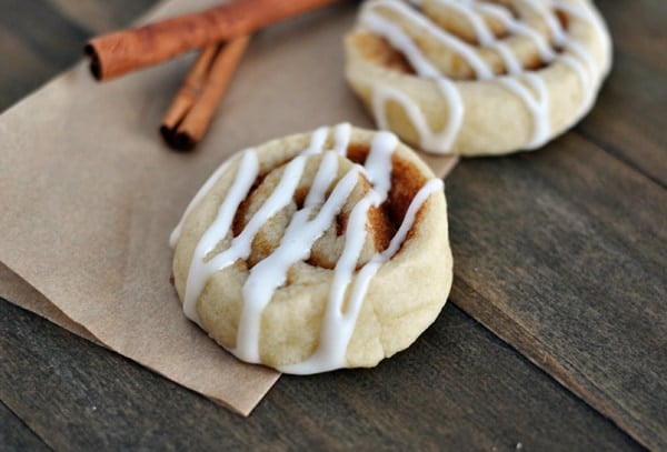 Two cinnamon roll sugar cookies with drizzled frosting on top next to a few cinnamon sticks.