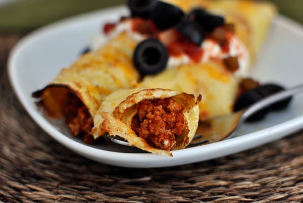 A fork taking a bite of taco meat filled cornmeal crepe topped with black olives, salsa, and sour cream.