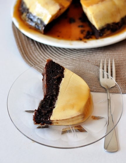 A piece of chocolate flan cake on a glass plate with the rest of the cake in the background.