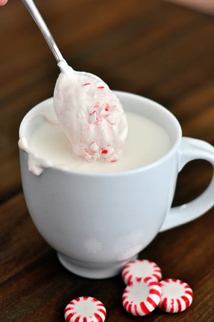 A white mug filled with white hot chocolate and a spoon with white chocolate and crushed peppermint coming out of the cup.