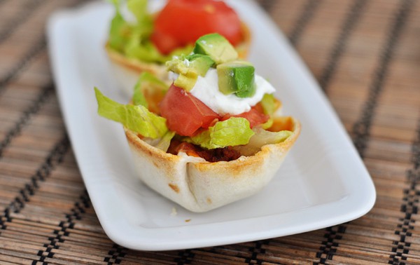 two mini taco cups topped with lettuce, tomato, sour cream, and diced avocado on a white platter