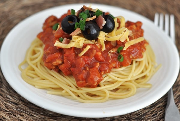A white plate with cooked spaghetti topped with red sauce, shredded cheese, and black olives.